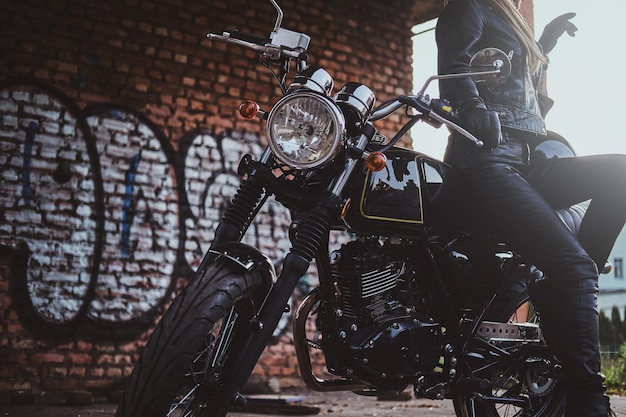 Stylowa seksowna kobieta w stroju motocyklisty pozuje dla fotografa obok swojego roweru i ściany z graffiti.