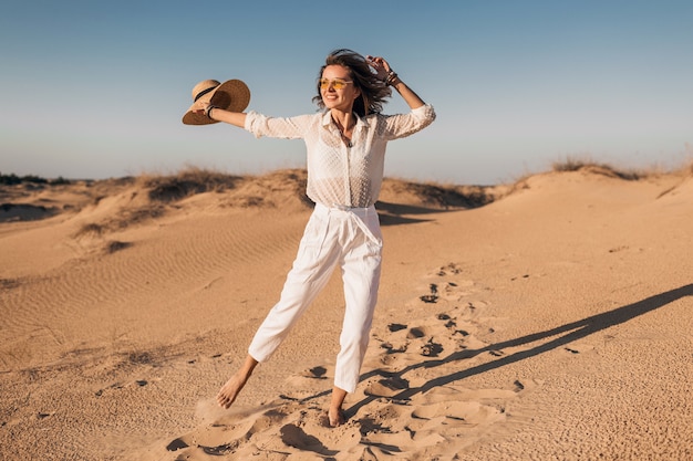 Stylowa uśmiechnięta piękna szczęśliwa kobieta bieganie i skakanie w piasku pustyni w białym stroju na sobie słomkowy kapelusz na zachód słońca