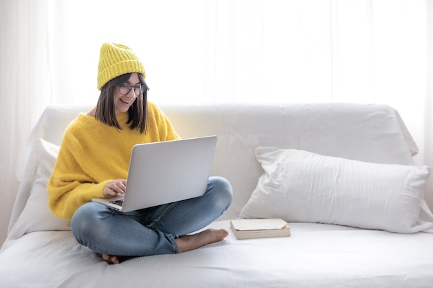 Bezpłatne zdjęcie stylowa wesoła brunetka dziewczyna w okularach i kapeluszu siedzi na kanapie w domu i pracuje na laptopie