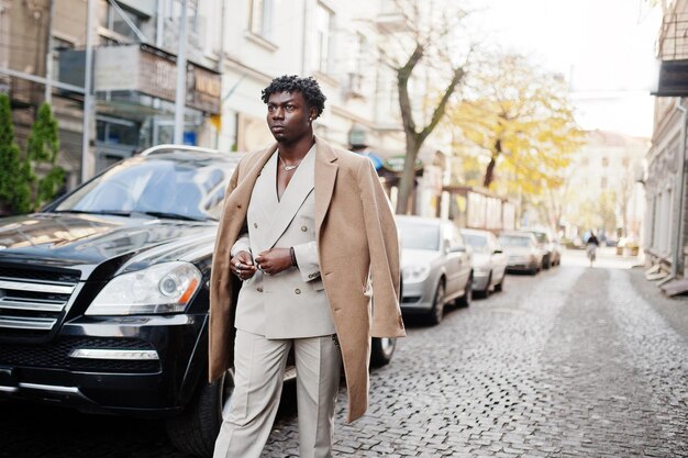 Stylowy afro mężczyzna w beżowym starym szkolnym garniturze i płaszczu chodzącym przed czarnym biznesowym samochodem Modny młody afrykański mężczyzna w casualowej kurtce na nagim torsie
