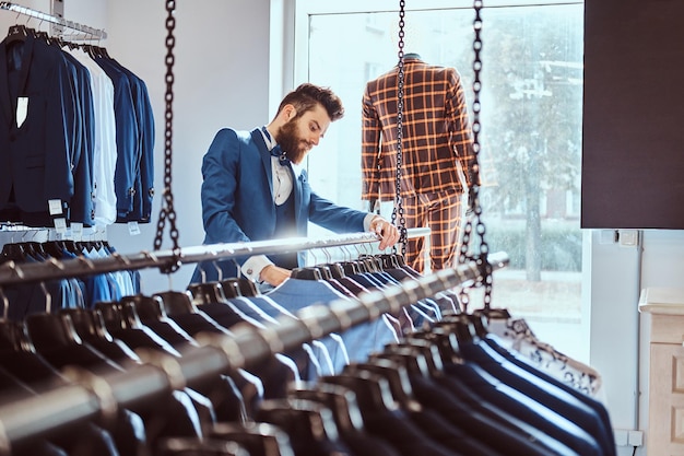 Stylowy brodaty sprzedawca ubrany w niebieski elegancki garnitur pracujący w sklepie z odzieżą męską.