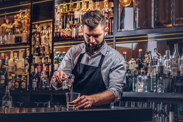 Stylowy brutalny barman w koszuli i fartuchu robi koktajl na tle baru.