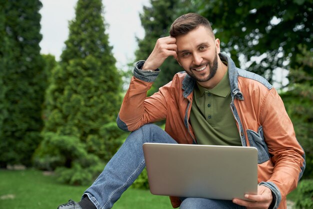 Stylowy mężczyzna pozuje siedząc z laptopem w ogrodzie