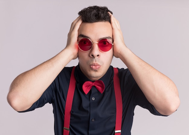 Stylowy mężczyzna z muszką w okularach i na szelkach patrząc z przodu zdezorientowany i zmartwiony, stojąc nad białą ścianą z rękami na głowie
