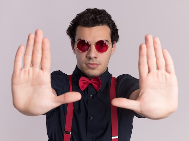 Bezpłatne zdjęcie stylowy mężczyzna z muszką w okularach i szelkach patrząc z przodu z poważną twarzą pokazującą gest stopu z rękami stojącymi na białej ścianie