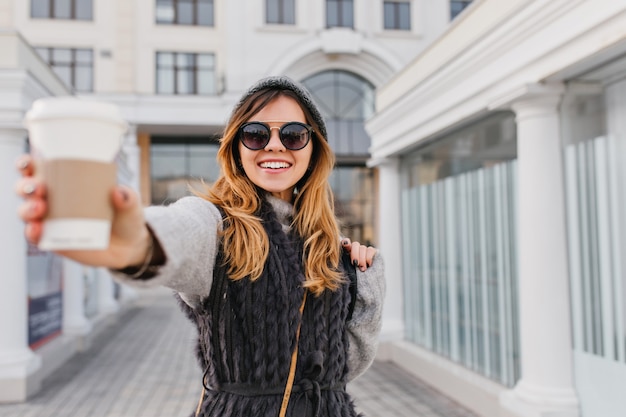 Stylowy portret miasta uśmiechnięta, radosna młoda kobieta, rozciąganie kawy na wynos. Dzień dobry modnej kobiety w nowoczesnych okularach przeciwsłonecznych, wełnianym swetrze, zabawy na świeżym powietrzu.