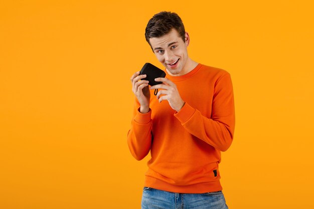 Stylowy uśmiechnięty młody człowiek w pomarańczowym swetrze trzymając bezprzewodowy głośnik szczęśliwy słuchając muzyki