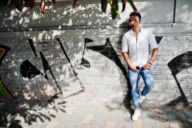 Stylowy wysoki arabski model człowieka w białej koszuli dżinsach i okularach przeciwsłonecznych postawiony na ulicy miasta Broda atrakcyjny arabski facet na ścianie graffiti