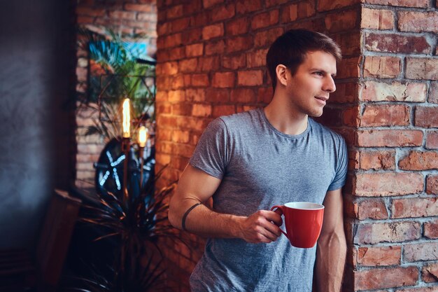 Stylowy, wytatuowany, atrakcyjny mężczyzna, ubrany w dżinsy i koszulkę, opiera się o ceglaną ścianę, trzymając w dłoni filiżankę kawy. Uśmiechając się i odwracając wzrok.