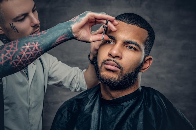 Stylowy wytatuowany fryzjer robi fryzurę czarnemu brodatemu mężczyźnie.