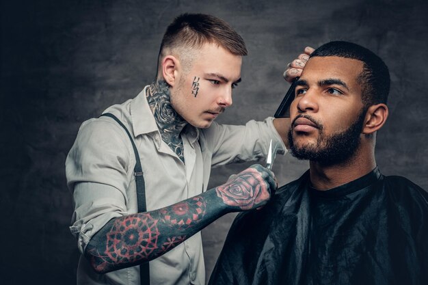 Stylowy wytatuowany fryzjer robi fryzurę czarnemu brodatemu mężczyźnie.