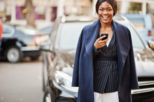 Sukces stylowej afroamerykańskiej kobiety w płaszczu przeciwko czarnemu biznesowemu samochodowi suv patrzącemu na telefon