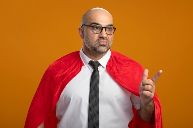 Superbohater biznesmen w czerwonej pelerynie i okularach patrząc na bok z poważną twarzą wskazującą palcem wskazującym na coś stojącego nad pomarańczową ścianą