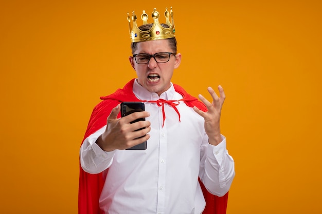 Bezpłatne zdjęcie superbohater biznesmen w czerwonej pelerynie i okularach w koronie trzymającej smartfona, patrząc na to z podniesioną ręką, zły i sfrustrowany, stojąc na pomarańczowym tle