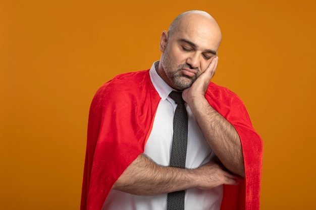 Superbohater biznesmen w czerwonej pelerynie, patrząc z przodu ze smutnym wyrazem twarzy, opierając głowę na dłoni zmęczony i znudzony stojąc nad pomarańczową ścianą