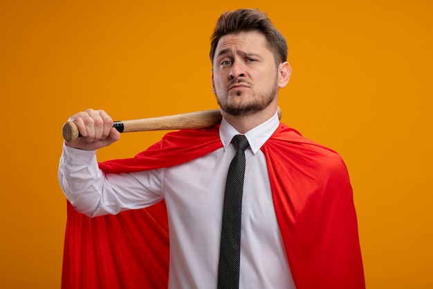 Superbohater biznesmen w czerwonej pelerynie trzyma kij baseballowy na ramieniu patrząc pewnie stojąc nad pomarańczową ścianą