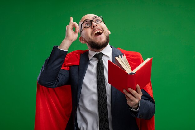 Superbohater Brodaty biznesmen w czerwonej pelerynie trzyma otwartą książkę patrząc w górę pokazując palec wskazujący mając pomysł uśmiechnięty stojący nad zieloną ścianą