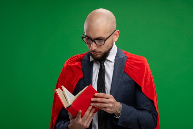 Superbohater Brodaty biznesmen w czerwonej pelerynie trzymając otwartą książkę czytania stojąc nad zieloną ścianą