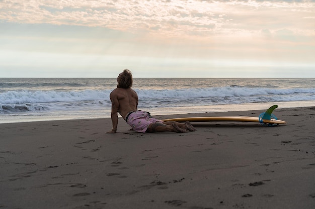 Surfer idzie z deską po piaszczystej plaży. Przystojny młody człowiek na plaży. Sporty wodne. Zdrowy, aktywny tryb życia. Surfing. Letni wypoczynek. Sport ekstremalny.