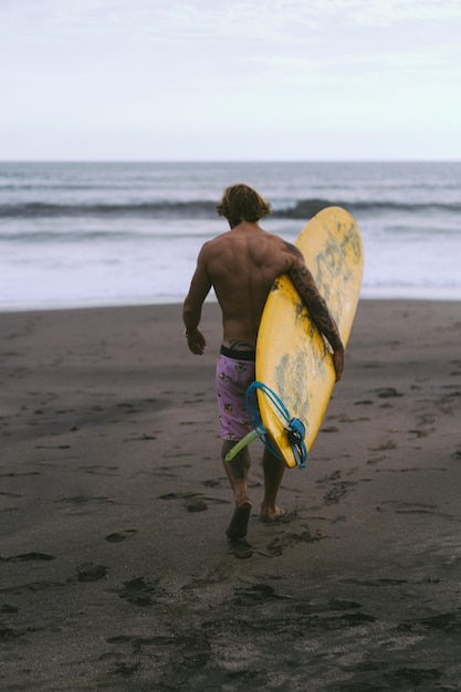 Surfer idzie z deską po piaszczystej plaży. Przystojny młody mężczyzna na plaży. Sporty wodne. Zdrowy, aktywny tryb życia. Surfing. Letni wypoczynek. Sport ekstremalny.
