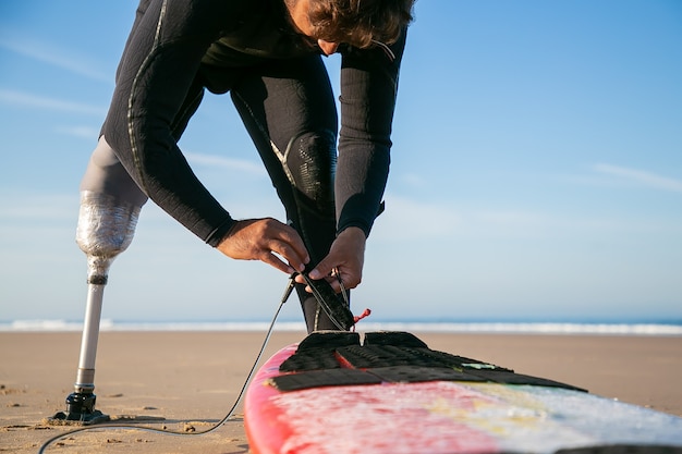 Bezpłatne zdjęcie surfer mężczyzna ubrany w piankę i sztuczną kończynę, przywiązujący deskę surfingową do kostki na piasku