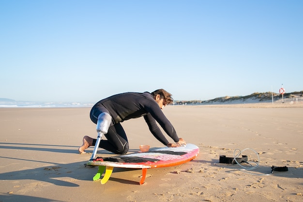 Surfer w piance w sztucznej kończynie, woskowany deskę surfingową na piasku na plaży oceanu