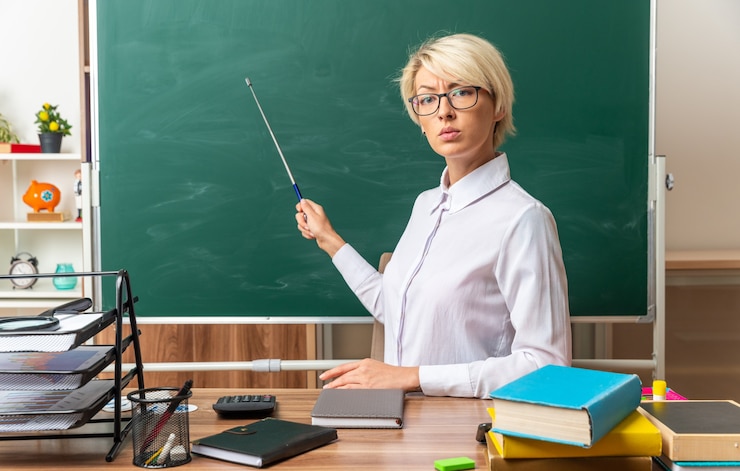 Bezpłatny plik Zdjęcie surowa młoda blond nauczycielka w okularach siedzi przy biurku z przyborami szkolnymi w klasie, wskazując na tablicę kijem wskaźnikowym, patrząc na przód