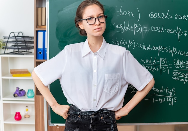 Surowa Młoda Blondynka Nauczycielka Matematyki W Okularach, Stojąc Przed Tablicą, Trzymając Ręce W Talii, Patrząc Na Kamerę W Klasie