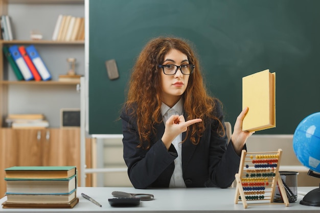 surowa młoda nauczycielka w okularach trzymająca i wskazująca na książkę siedząca przy biurku z szkolnymi narzędziami w klasie