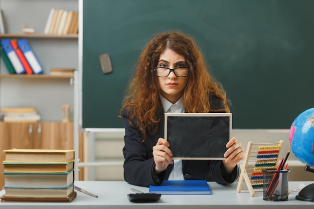 Bezpłatne zdjęcie surowa młoda nauczycielka w okularach trzymająca mini tablicę siedzącą przy biurku z szkolnymi narzędziami w klasie