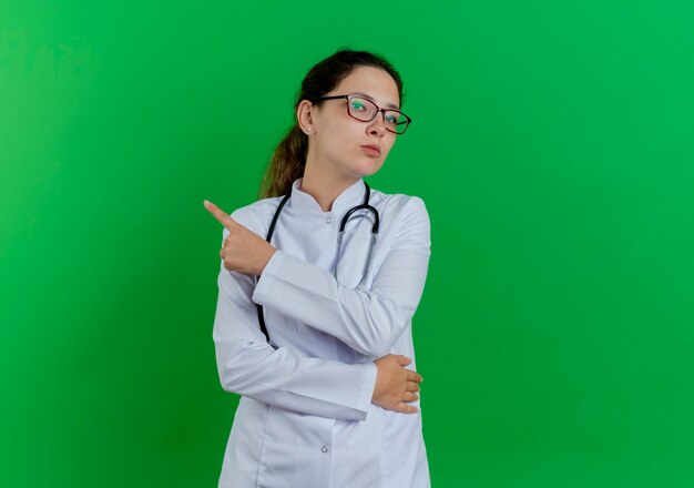 Surowe młoda kobieta lekarz ubrana w szlafrok medyczny i stetoskop i okulary, wskazując na bok na białym tle na zielonej ścianie z miejsca na kopię
