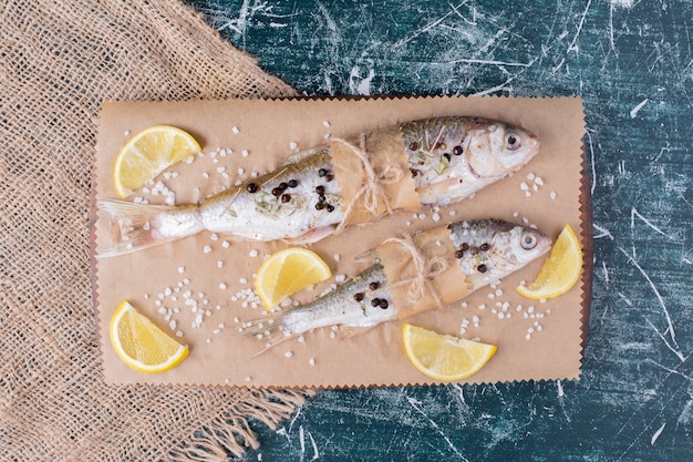 Bezpłatne zdjęcie surowe ryby całe z plasterkami cytryny, ziarnami pieprzu i solą na desce.