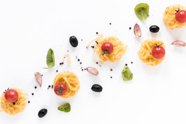 Surowy makaron fettuccine; pomidor wiśniowy; czarna oliwka; Ząbek czosnku i liści bazylii samodzielnie na białym tle