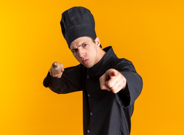 Surowy młody blond mężczyzna kucharz w mundurze szefa kuchni i czapce stojącej w widoku profilu, patrząc i wskazując na kamerę z wałkiem i palcem na białym tle na pomarańczowej ścianie