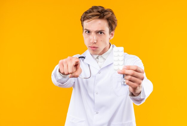 Surowy młody lekarz mężczyzna ubrany w szatę medyczną ze stetoskopem trzymającym pigułki pokazujące gest
