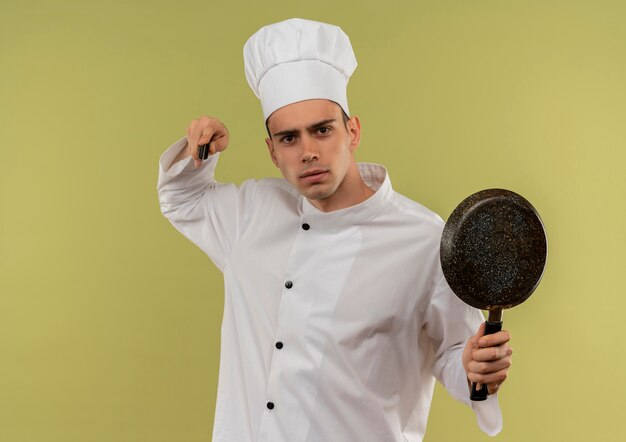 Surowy młody mężczyzna kucharz ubrany w mundur szefa kuchni trzymając patelnię podnosząc nóż w ręku