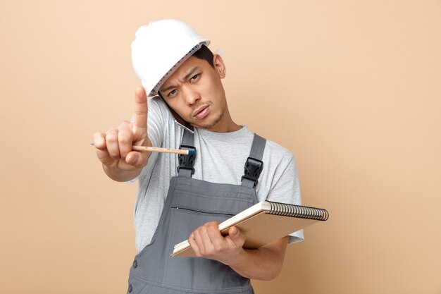 Surowy młody pracownik budowlany w kasku ochronnym i mundurze trzymającym notatnik i ołówek robi trzymający gest