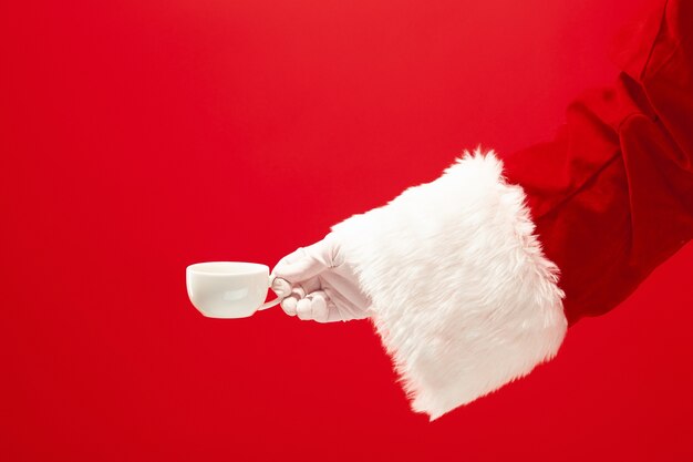 Świąteczna kawa. Santa ręka trzyma filiżankę kawy na białym tle na czerwonym tle z miejscem na tekst.
