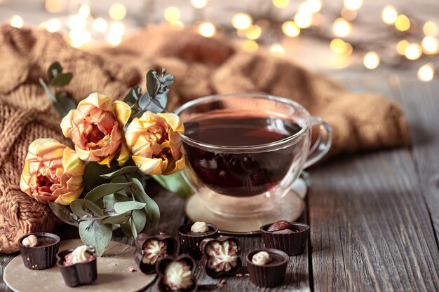 Świąteczna martwa natura z drinkiem w filiżance, czekoladki i kwiaty na niewyraźnym tle z bokeh.