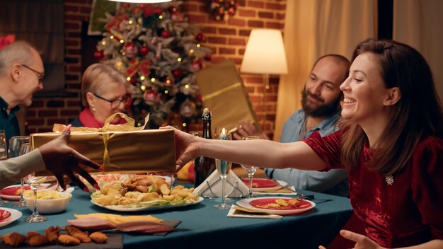Świąteczni członkowie rodziny siedzą przy świątecznym stole podczas wymiany prezentów w domu. Radosni ludzie zebrali się w salonie, aby świętować zimowe tradycyjne święta, jednocześnie obdarowując się prezentami.