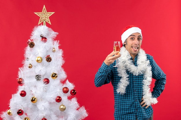 Bezpłatne zdjęcie Świąteczny nastrój z emocjonalnym młodzieńcem w kapeluszu świętego mikołaja i podnosząc kieliszek wina wiwatuje w pobliżu choinki