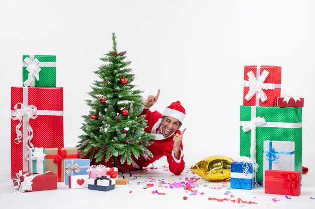 Świąteczny nastrój z młodym zabawnym mikołajem chowającym się za choinką w pobliżu prezentów w różnych kolorach na białym tle