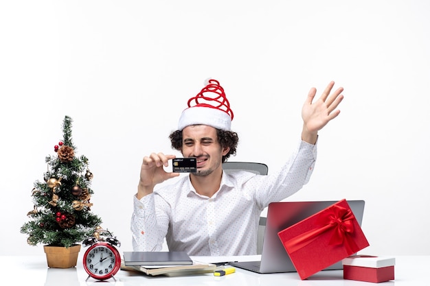Świąteczny nastrój z młodym zmęczonym, gniewnym biznesmenem w kapeluszu świętego mikołaja i patrząc na swoją kartę bankową w biurze na białym tle