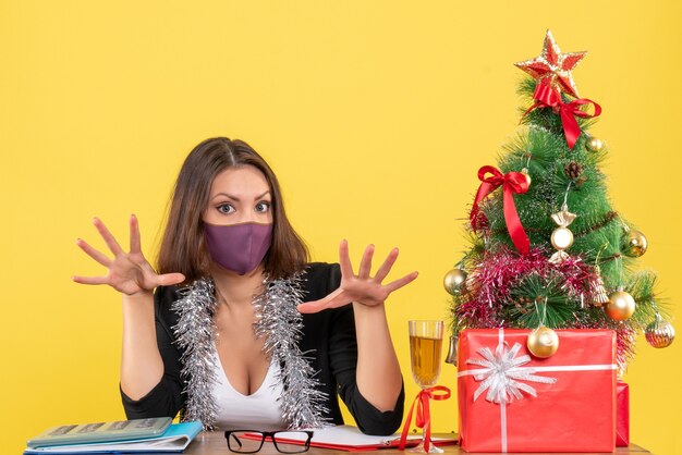 Świąteczny nastrój z uroczą damą w garniturze na sobie maskę medyczną w biurze na żółto na białym tle