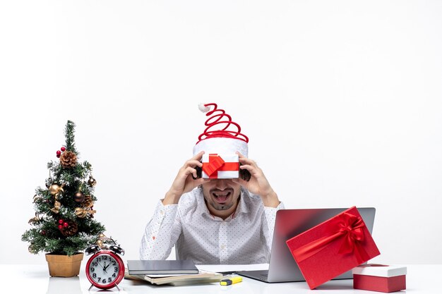 Świąteczny nastrój z zabawnym biznesmenem w czapce świętego mikołaja trzymającego prezent przed twarzą i wystawiającego język na białym tle