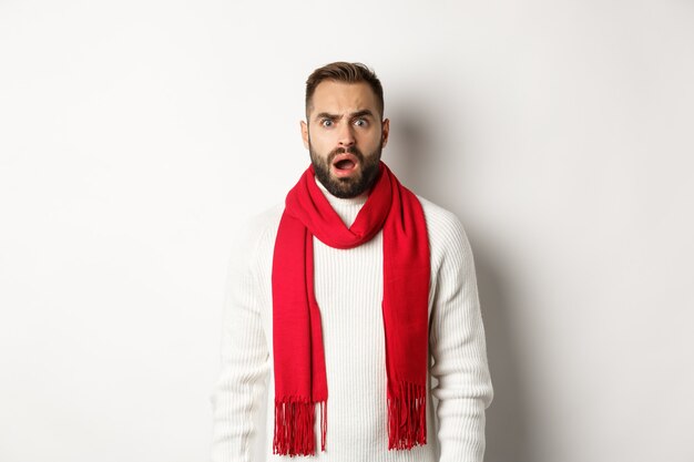 Święta Bożego Narodzenia i koncepcja uroczystości. Zdezorientowany brodaty facet wpatrujący się w coś dziwnego, stojący w czerwonym szaliku i swetrze, białe tło.