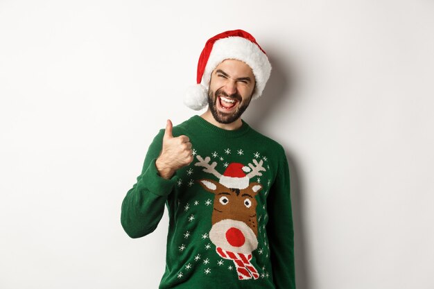 Święta Bożego Narodzenia, koncepcja uroczystości i partii. Mężczyzna cieszy się Nowy Rok, pokazując kciuk do góry w aprobacie, ubrany w Santa hat, białe tło.