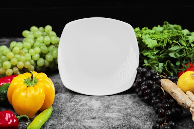 Bezpłatne zdjęcie Świeże warzywa, winogrona, papryka, zieleń, cytryna, pomidor i biały talerz na ciemnym tle.