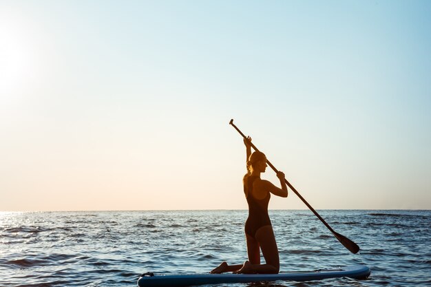 Sylwetka młody piękny kobieta surfing w morzu przy wschodem słońca.