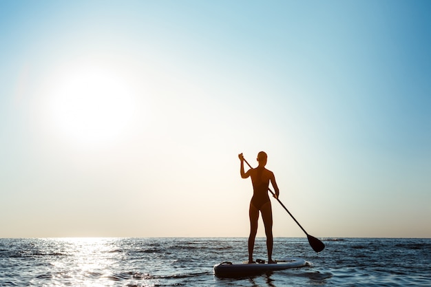 Sylwetka młody piękny kobieta surfing w morzu przy wschodem słońca.